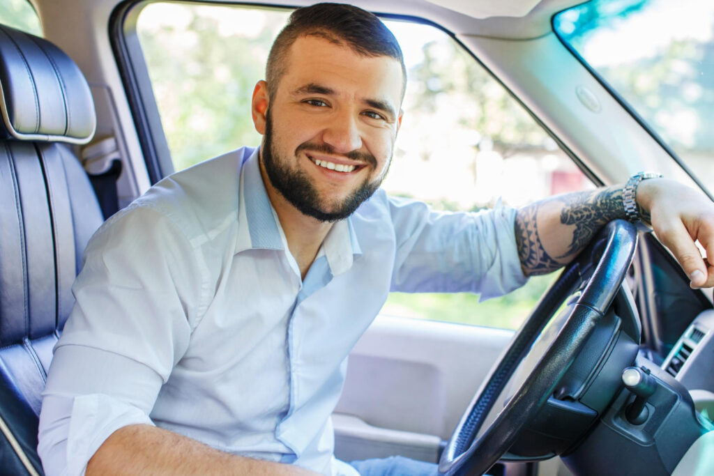 happy male with black hair beard tattoes his hand dressed white shirt blue jeans shorts driving car 1